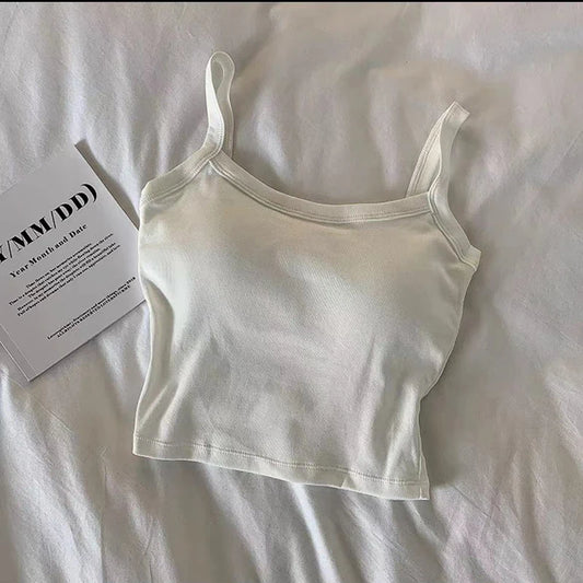 The Crop Top Women Sexy Sleeveless Tops in white, featuring thin straps and a cropped fit, is laid flat on a white bedsheet next to an open booklet—perfect for effortless summer style.